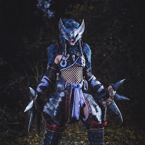 Puede incluir: Un cosplayer vestido con una armadura azul y negra con un casco azul y plateado. El traje incluye una faja morada y un par de cuchillas plateadas. El cosplayer est&aacute; de pie en un entorno de bosque.