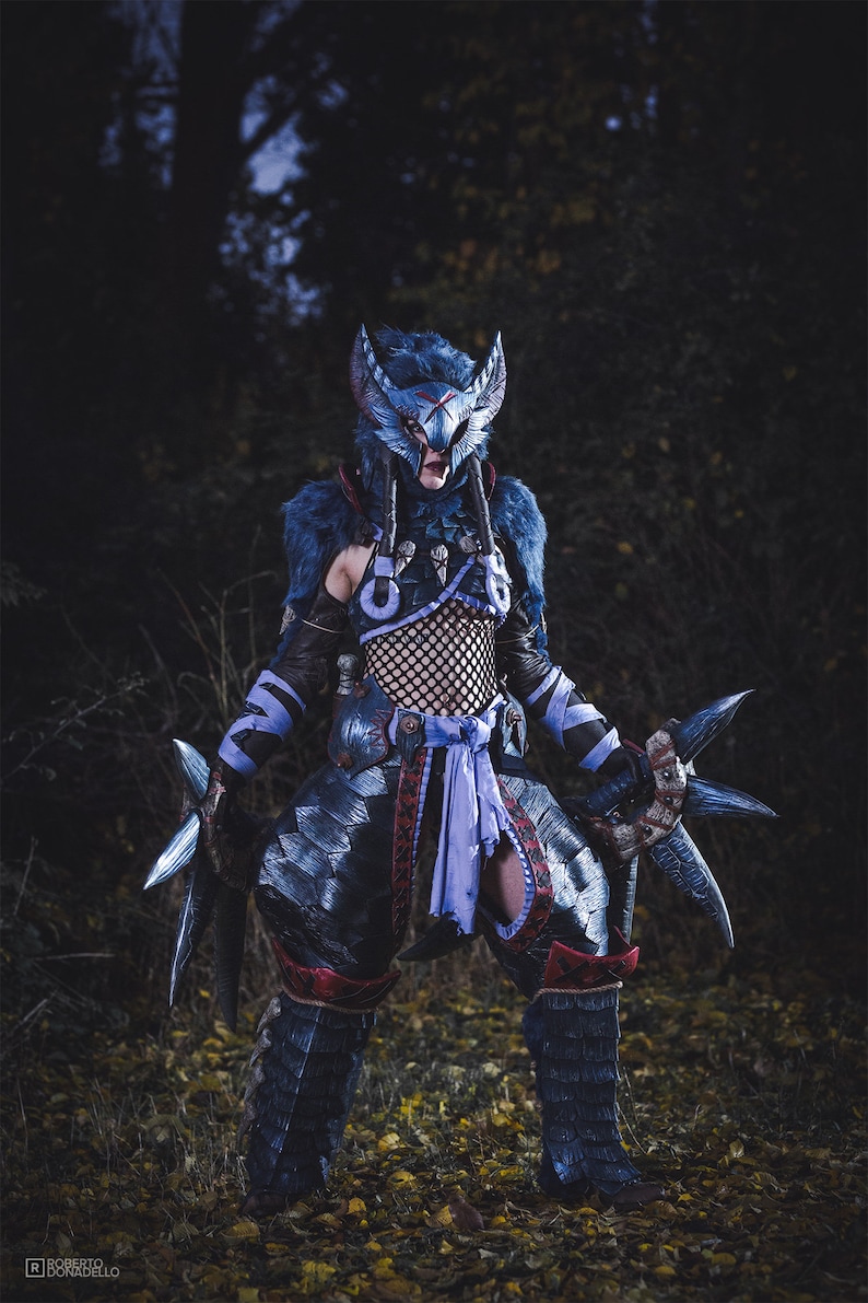 Puede incluir: Un cosplayer vestido con una armadura azul y negra con un casco azul y plateado. El traje incluye una faja morada y un par de cuchillas plateadas. El cosplayer est&aacute; de pie en un entorno de bosque.