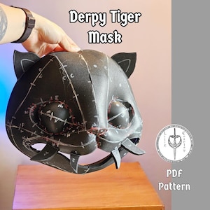 Puede incluir: Una máscara de tigre negra con el texto "Derpy Tiger Mask" es sostenida por una mano. La máscara tiene marcas blancas y está hecha de piezas segmentadas. La imagen también incluye un logotipo y las palabras "PDF Pattern".