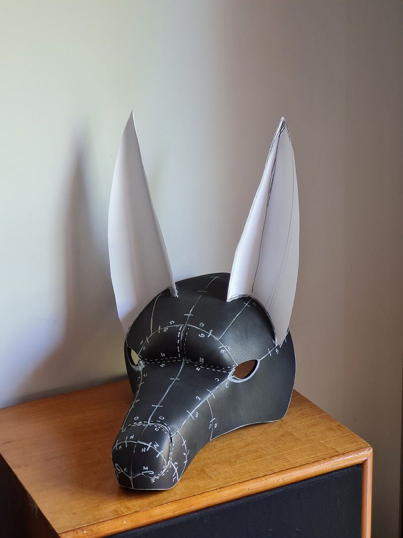 May include: A black and white furry mask with large ears. The mask is made of foam and has a pattern of lines and numbers on it. The mask is sitting on a wooden surface.