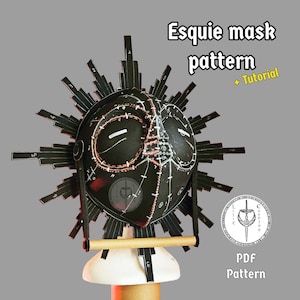 Könnte beinhalten: Eine schwarze Maske mit einem Sonnenstrahl-Design, mit weißen und roten Akzenten. Die Maske hat ein einzigartiges, geometrisches Muster und ist auf einem Ständer montiert. Der Text auf dem Bild lautet "Esquie mask pattern + Tutorial" und "PDF Pattern".