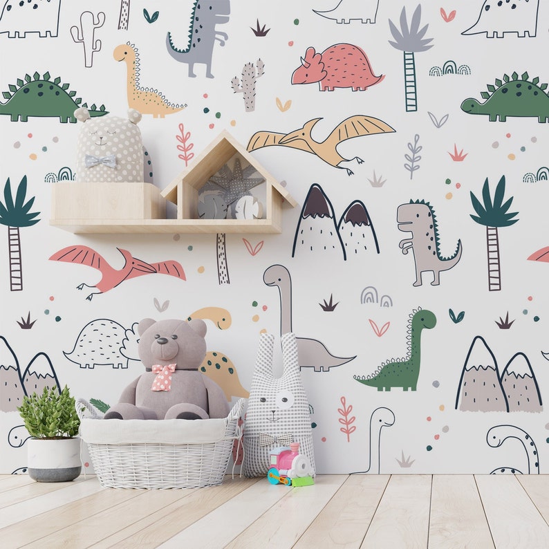 May include: A white wall with a dinosaur themed wallpaper featuring various dinosaurs, mountains, and plants. A wooden shelf with a stuffed animal on top. A teddy bear sitting in a white wicker basket. A gray bunny stuffed animal on the floor.