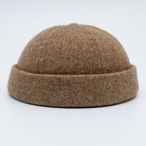 Puede incluir: Un gorro marrón, tipo beanie, hecho de material similar a la lana, con un borde doblado. El gorro tiene una corona redondeada y una superficie texturizada. El color es un marrón cálido y terroso, adecuado para el clima frío. Un estilo clásico.