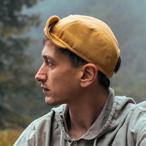 Könnte beinhalten: Eine Person mit einer senffarbenen Kappe und einem hellgrauen Kapuzenshirt sitzt auf einer Straße. Der Hintergrund zeigt einen nebligen Wald und eine kurvenreiche Straße, was eine ruhige Szene im Freien schafft.