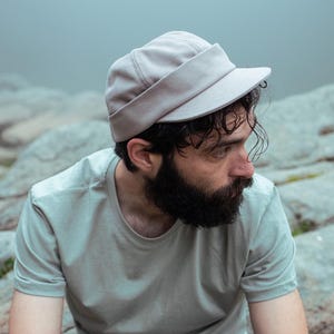 May include: A person wearing a light grey cap and a light green t-shirt sits on a rocky surface. The cap has a short brim and a band around the head. The t-shirt is short-sleeved. The background is a misty, outdoor scene.