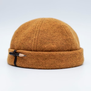 Puede incluir: Gorro de color mostaza con un lápiz adjunto. El gorro está hecho de un material suave y texturizado y presenta un borde doblado. Una correa negra sujeta un lápiz en su lugar, agregando un detalle único al diseño del gorro. El gorro es un accesorio elegante.