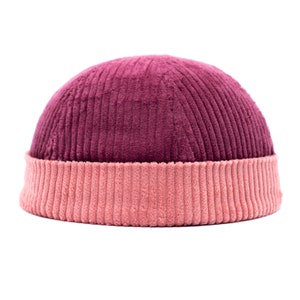 May include: A two-tone corduroy beanie hat. The crown of the hat is a deep burgundy colour, while the folded brim is a lighter, dusty rose colour. The hat has a ribbed texture.