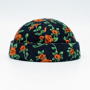 May include: A navy blue beanie hat with a floral pattern of orange roses and green leaves. The hat is made of a corduroy fabric and has a rolled-up brim. The hat is a stylish accessory.