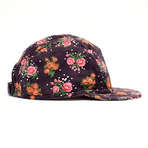May include: A dark purple corduroy five-panel hat with a floral pattern. The flowers are pink and orange with white accents. The hat has a flat brim and a strap in the back for adjustment.