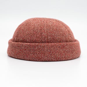 Könnte beinhalten: Eine rotbraune, strukturierte Beanie-Mütze mit umgeschlagenem Rand. Die Mütze besteht aus einem gewebten Stoff und liegt auf einer weißen Oberfläche. Die Farbe der Mütze ist eine Mischung aus roten und braunen Fäden.