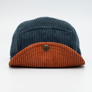 May include: A corduroy cap in a dark teal color with a contrasting burnt orange brim. The cap features a button detail at the front and a classic five-panel design. The fabric has a ribbed texture.