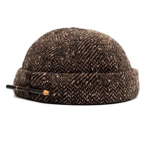 May include: A brown tweed cap with a herringbone pattern. The cap features a leather band with two pencils attached. The hat is a dome shape and is set against a white background.