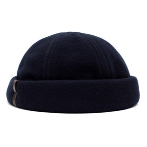 Könnte beinhalten: Eine marineblaue Beanie-Mütze mit umgeschlagenem Saum. Die Mütze hat ein strukturiertes Gewebe und einen kleinen braunen Lederakzent an der Seite. Die Mütze ist so konzipiert, dass sie eng am Kopf anliegt.