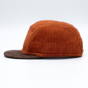 Op de afbeelding: Bruine corduroy baseballcap met een platte klep. De pet heeft een warme, roodbruine kleur, en de klep is een donkerdere bruintint. De pet heeft een klassiek vijf-panelen ontwerp.