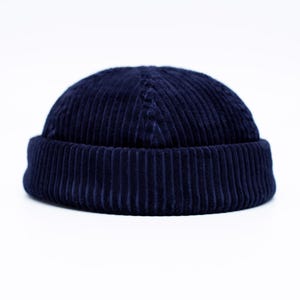 Puede incluir: Un gorro beanie de pana azul marino. El gorro tiene una textura acanalada y un borde doblado. El gorro es de color liso y está sobre un fondo blanco. Este gorro es un accesorio elegante para el clima frío.