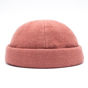 Può includere: Un berretto color rosa antico con bordo arrotolato. Il cappello è realizzato in un tessuto strutturato e si staglia su uno sfondo bianco. Il berretto è uno stile aderente, adatto all'abbigliamento casual.
