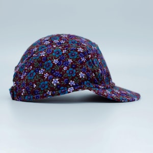 Op de afbeelding: Een baseballcap met een bloemenpatroon. De pet heeft een bordeauxrode achtergrond met een herhalend patroon van blauwe en paarse bloemen. De klep is gebogen en de pet is gemaakt van een textuurstof.