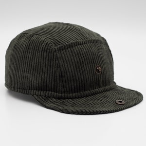 May include: A dark green corduroy cap with a short brim. The cap has a structured design with a button detail on the side and a metal eyelet on the brim. The fabric has a ribbed texture.