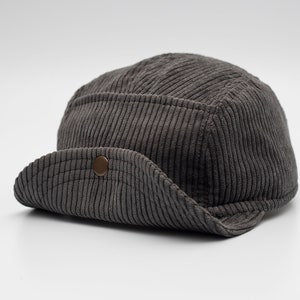 May include: A grey corduroy cap with a rounded crown and a curved peak. The cap has a button detail on the peak and a textured, ribbed fabric. The hat is a neutral colour and suitable for various outfits.