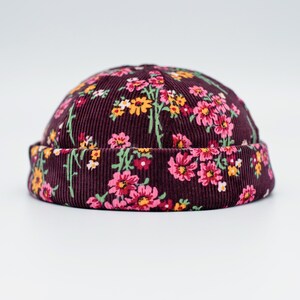 Op de afbeelding: Een bordeauxrode corduroy beanie-muts met een bloemenpatroon. De hoed heeft een herhalend patroon van roze, gele en witte bloemen met groene stelen en bladeren. De hoed heeft een afgeronde vorm met een omgeslagen rand.