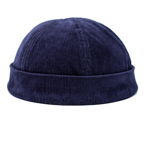 Può includere: Un berretto in velluto a coste blu navy con bordo arrotolato. Il cappello ha una corona arrotondata e una trama a coste. Il cappello è realizzato in un tessuto morbido e felpato ed è adatto al clima freddo.