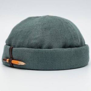 May include: A teal-coloured, rolled-brim beanie hat with a brown leather strap and a pencil attached. The hat is made of a textured fabric. The pencil is orange and brown.