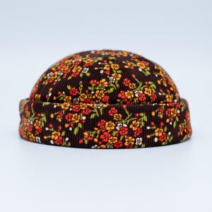 Könnte beinhalten: Ein brauner Cord-Beanie-Hut mit einem Blumenmuster in Rot-, Orange-, Gelb- und Weißtönen. Der Hut hat einen umgeschlagenen Rand und eine runde Krone. Das florale Design bedeckt den gesamten Hut.