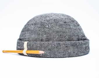 May include: A black and white herringbone patterned beanie hat with a rolled brim. A yellow pencil is attached to the hat with a cream-colored band. The hat is made of a textured fabric and sits on a white background.
