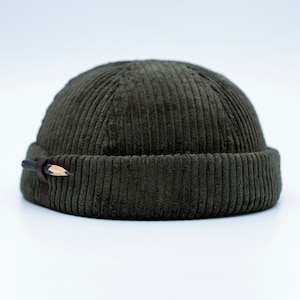 Puede incluir: Gorro beanie de pana verde oliva oscuro con un detalle de lápiz y correa de cuero. El gorro tiene una textura acanalada y un borde doblado. El lápiz está sujeto al lateral del gorro con una correa de cuero marrón.