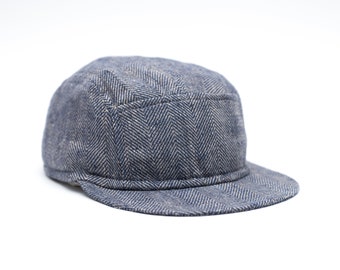 May include: A blue and gray herringbone pattern cap with a short brim. The cap has a rounded crown and a structured design. The fabric appears to be a textured tweed or similar material, suitable for cooler weather. The cap is set against a white background.