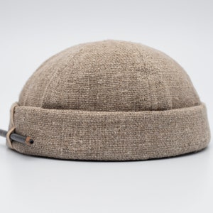 Pode incluir: Um gorro bege feito de um tecido texturizado. O chapéu tem uma aba enrolada e um pequeno lápis preso na lateral. O chapéu é de cor neutra e parece ser feito de um material tecido.