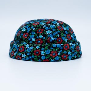 Könnte beinhalten: Eine Beanie-Mütze mit floralem Muster. Die Mütze hat einen schwarzen Hintergrund mit einem Muster aus roten und blauen Blumen. Der Rand der Mütze ist hochgekrempelt. Die Mütze ist aus einem weichen, strukturierten Stoff.