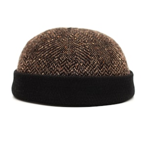 Può includere: Un berretto beanie con motivo a spina di pesce marrone e nero. Il berretto ha un bordo a coste nero e una corona in stile tweed. Uno stile classico, adatto al clima freddo.