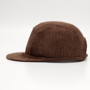 Könnte beinhalten: Eine braune Cord-Cap mit flachem Schirm. Die Kappe hat ein Fünf-Panel-Design und eine strukturierte Krone. Der Stoff hat eine vertikale Rippenstruktur. Ein neutrales Accessoire.