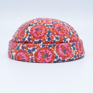 Peut inclure: Un bonnet rose avec un motif floral orange, bleu et blanc. Le chapeau est fait d'un tissu côtelé et a un bord roulé. Le motif floral est uniformément réparti sur la surface du chapeau.