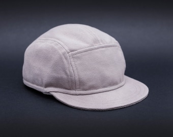 May include: A light gray, five-panel cap with a short brim. The cap is made of a textured fabric and features a structured design. The brim is curved, and the cap has a classic, casual style.