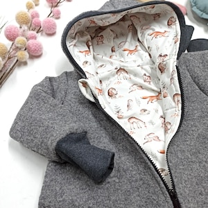 Könnte beinhalten: Eine graue Kapuzenjacke mit weißem Innenfutter, das mit einem wiederkehrenden Muster aus Waldtieren wie Füchsen, Rehen, Eulen und Hasen bedruckt ist. Die Jacke hat einen schwarzen Frontreißverschluss und dunkelgraue gerippte Bündchen an den Ärmeln.