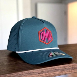 Peut inclure: Casquette de baseball bleu sarcelle avec un détail de corde blanche et une visière incurvée. Le chapeau présente un écusson hexagonal rose et rouge avec les lettres "OMB".