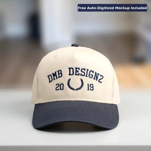 May include: A cream-coloured baseball cap with a navy blue peak and button. The cap has navy blue embroidered text reading "DMB DESIGNZ 20 19", with a laurel wreath. The text "Free Auto-Digitized Mockup Included" is in the top right corner.