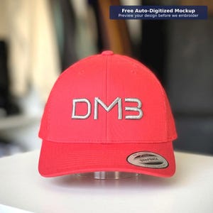 May include: A red baseball cap featuring the embroidered letters "DMB" in silver. The cap has a curved peak and a snapback closure. The text "Free Auto-Digitized Mockup Preview your design before we embroider" is in the top right corner.