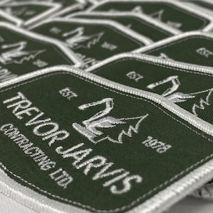 May include: A collection of green and white embroidered patches. Each patch displays the text "TREVOR JARVIS CONTRACTING LTD." and a graphic. The patches have a white border and the text "EST. 1978".