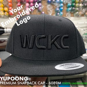 May include: Black and green baseball cap with the text "WCKC" embroidered in black on the front. The cap has a black adjustable strap with a silver buckle and a black under-visor. The text "YUPOONG® PREMIUM SNAPBACK CAP - 6089M (GREEN UNDERVISOR)" is printed on the bottom of the cap.