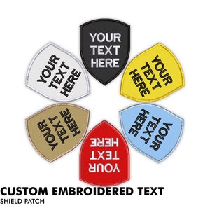 May include: Six embroidered shield patches in different colours: black, white, yellow, beige, red, and light blue. Each patch features the text "YOUR TEXT HERE" in black.