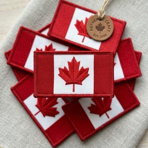 May include: Embroidered patches of the Canadian flag, featuring a red and white design with a maple leaf. A small, round leather tag with the text "AUTHENTIC CRAFT EST. 2024" is attached. Patches are arranged on a neutral linen surface.