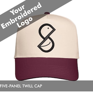 Peut inclure: Casquette en sergé à cinq panneaux avec un panneau avant crème et une visière bordeaux. La casquette présente un logo noir brodé sur le panneau avant. Le texte "Your Embroidered Logo" est au-dessus de la casquette.