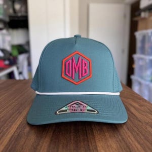 Peut inclure: Casquette de baseball bleu sarcelle avec un détail de corde blanche et une visière plate. Le chapeau présente un logo hexagonal brodé rose et orange avec les lettres "OMB". La visière porte l'inscription "PACIFIC BUILT FOR YOU".