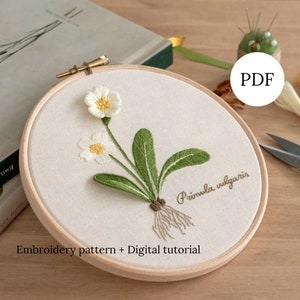 May include: Embroidered hoop art featuring a white primrose flower with green leaves and roots. The text "Primula vulgaris" is stitched below the flower. The hoop is on a wooden surface. The text "Embroidery pattern + Digital tutorial" is below the hoop. The word "PDF" is in a circle above the hoop.