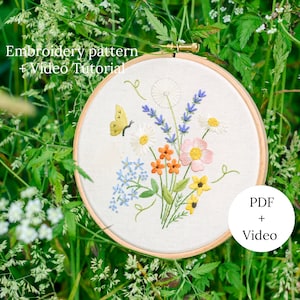 May include: Embroidered hoop art featuring a bouquet of colorful wildflowers, including daisies, lavender, and forget-me-nots, with a yellow butterfly. The hoop is surrounded by green grass. The text "Embroidery pattern + Video Tutorial" and "PDF + Video" are also visible.