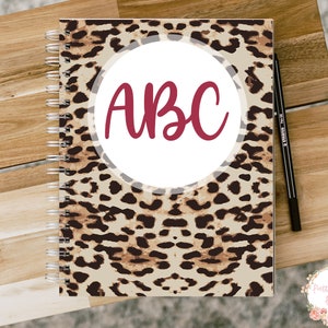 Pode incluir: Um caderno espiralado com uma capa de estampa de leopardo. A capa tem um c&iacute;rculo branco com as letras "ABC" em cor bord&ocirc;.