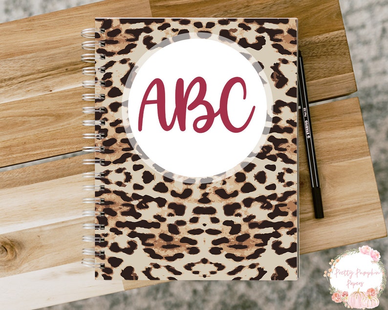 Pode incluir: Um caderno espiralado com uma capa de estampa de leopardo. A capa tem um c&iacute;rculo branco com as letras "ABC" em cor bord&ocirc;.